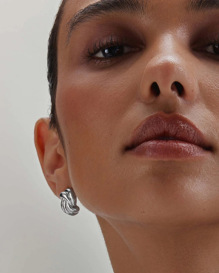 A close-up of a model's face wearing a small silver twisted knot stud earring against a plain light background.