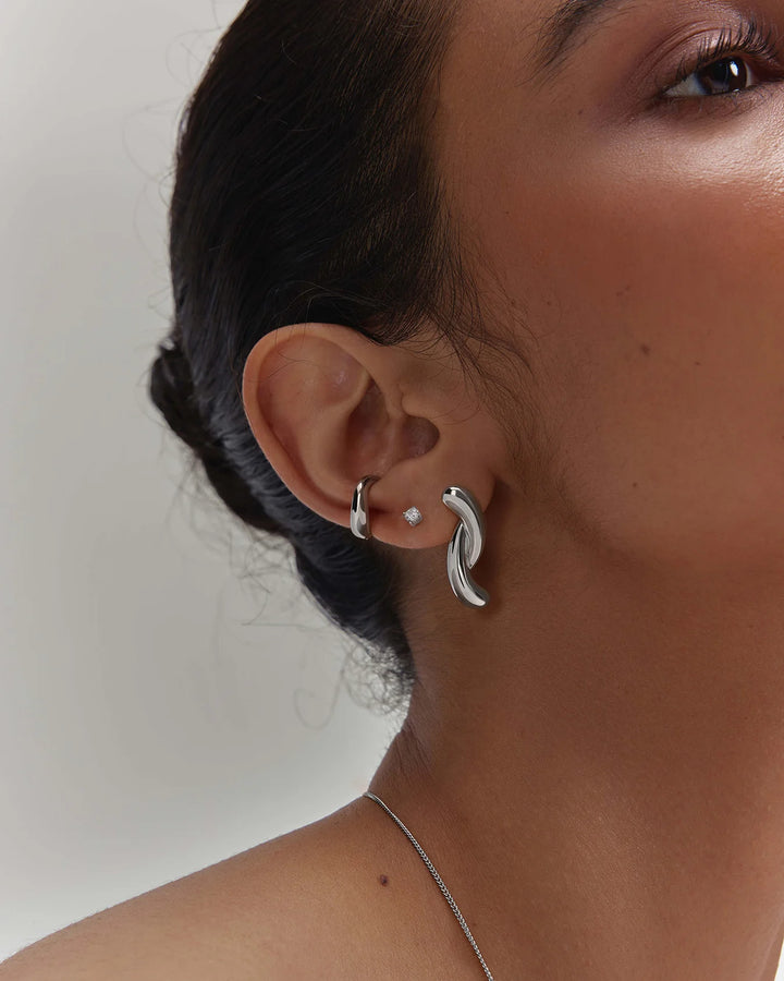 A close-up of a model's ear wearing a gold huggie hoop earring and a larger gold crescent-shaped dangle earring, with a small white pearl stud also visible.
