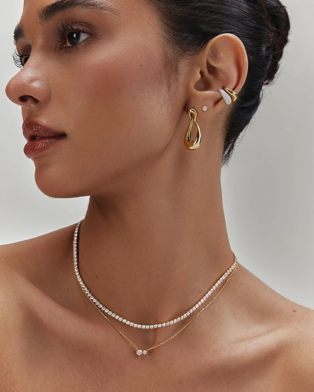 A close-up of a model wearing layered gold and pearl jewelry, including a delicate pearl necklace, a gold chain necklace with a small pearl pendant, a gold teardrop hoop earring, and a gold ear cuff with a small pearl stud.