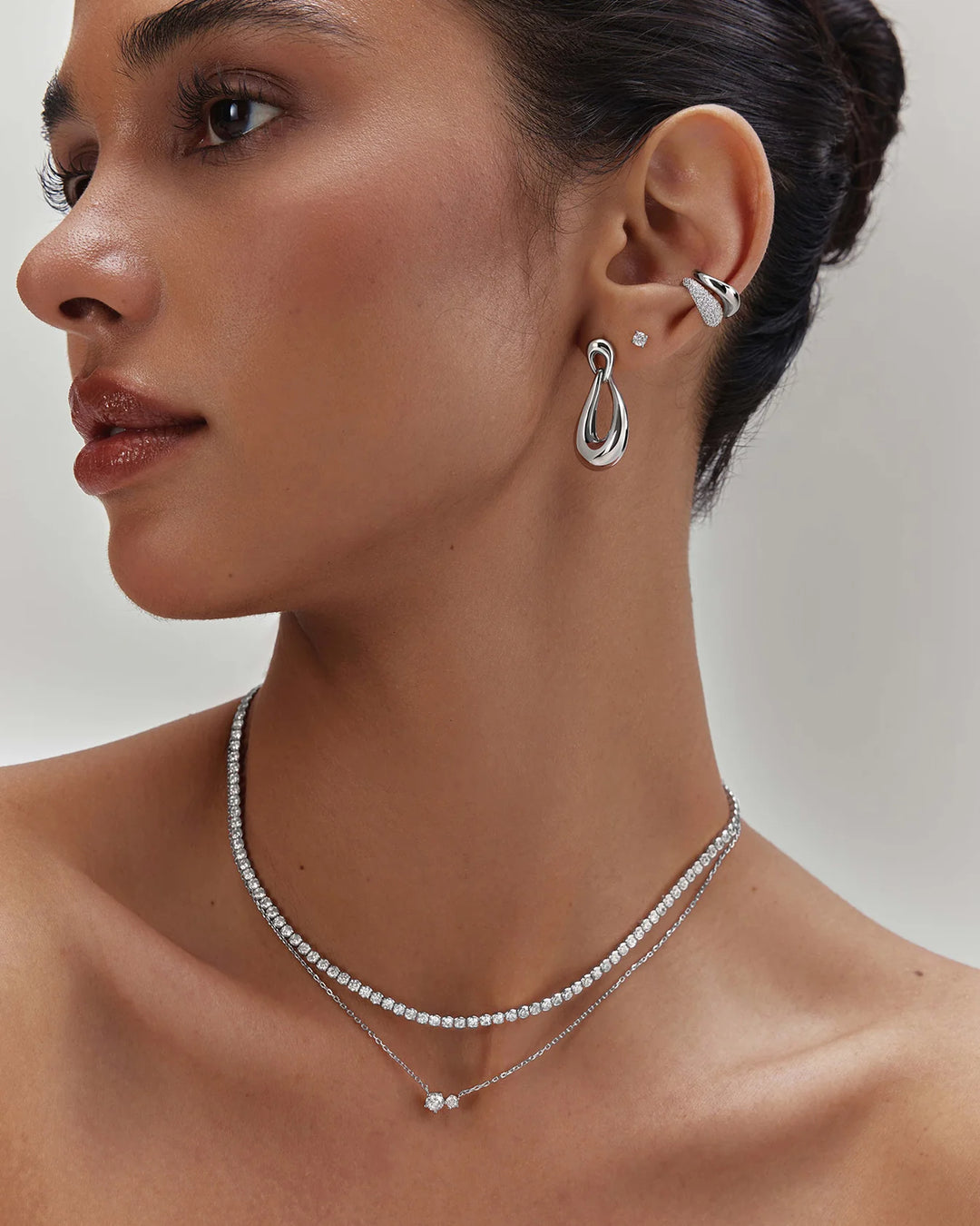 A close-up of a woman wearing a delicate silver double-layered necklace with a small pendant and a matching silver hoop earring with a small stud.