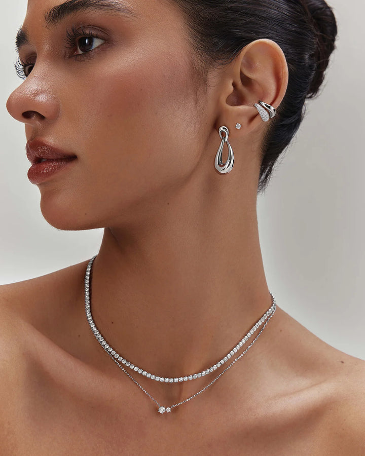 A close-up of a woman wearing a delicate silver double-layered necklace with a small pendant and a matching silver hoop earring with a small stud.