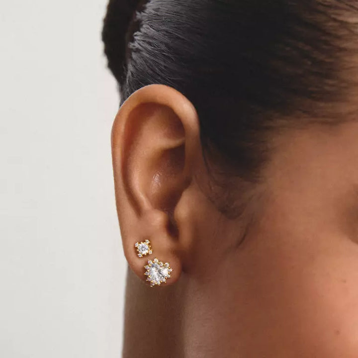 A close-up of a person's ear wearing a pair of gold floral stud earrings with clear gemstones, set against a plain light gray background.