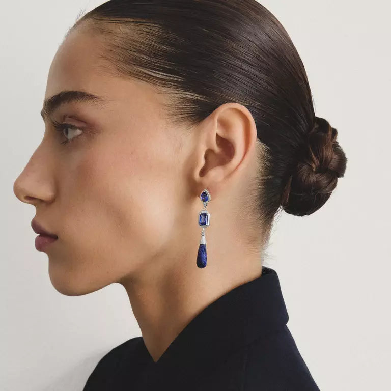 A close-up side profile of a woman with dark hair in a bun, wearing a single drop earring with deep blue stones and silver-toned metal details against a light gray background.