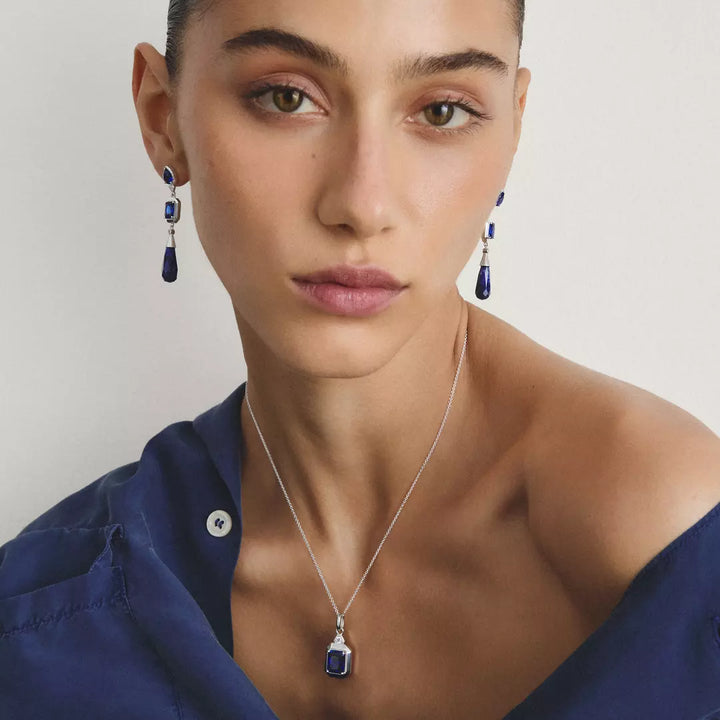 A close-up of a model wearing a silver necklace with a dark blue rectangular pendant and matching drop earrings, paired with a navy blue shirt against a plain light background.