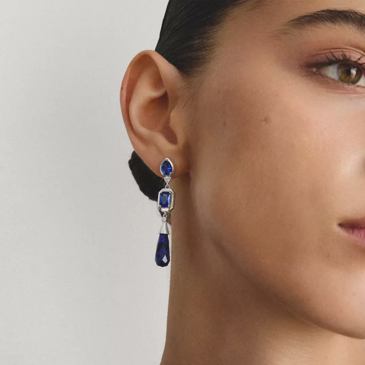 A close-up of a model's ear wearing a silver drop earring with deep blue gemstones.