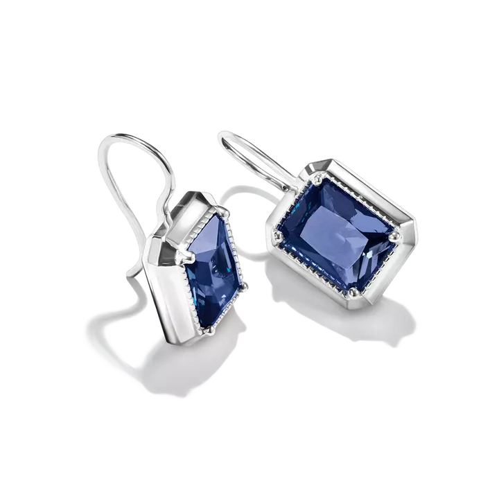 A pair of silver drop earrings featuring rectangular emerald-cut blue gemstones in silver bezel settings with small clear accent stones, shown on a white background.