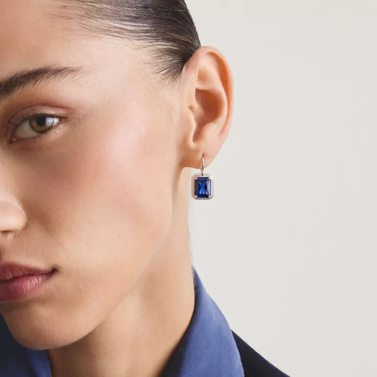 A close-up of a model's ear wearing a single drop earring with a rectangular deep blue gemstone in a silver-toned setting.