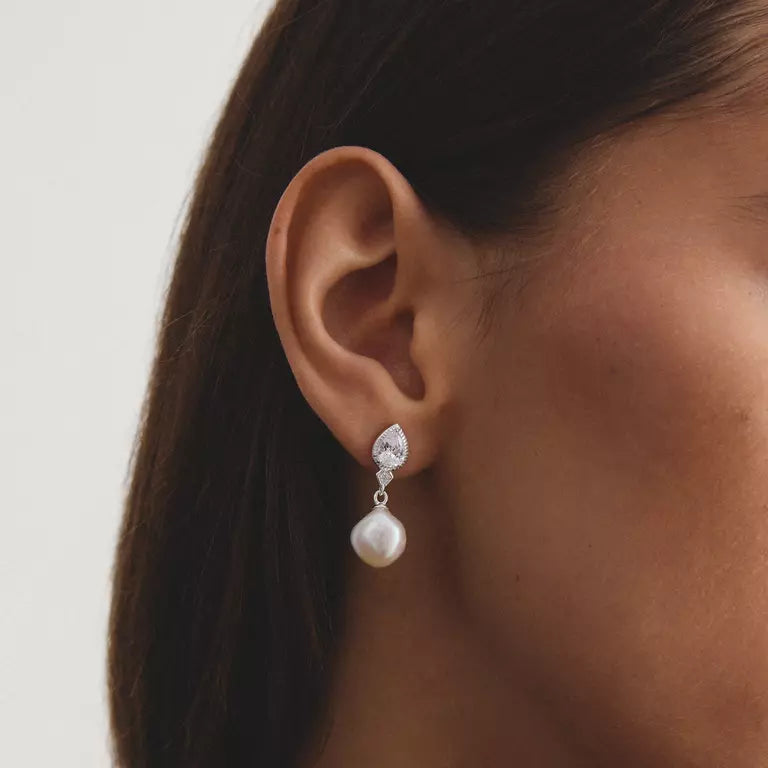 A close-up of a woman's ear wearing a silver drop earring with a clear teardrop-shaped stone and a dangling white pearl.