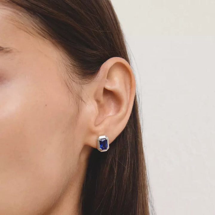 A close-up of a woman's ear wearing a small rectangular stud earring with a deep blue gemstone in a silver setting, against a plain light background.