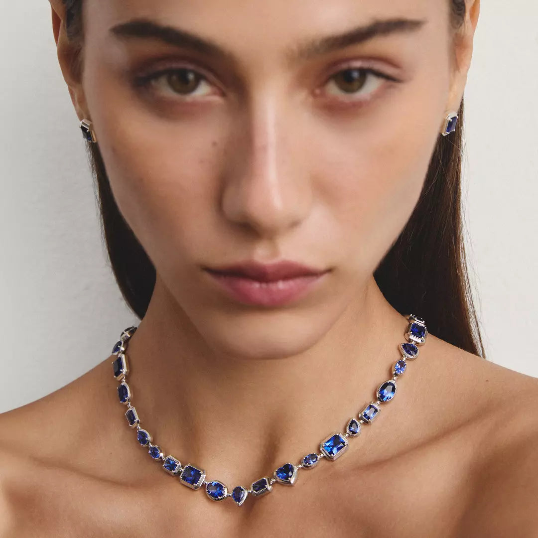A close-up of a model wearing a silver-toned necklace and matching stud earrings set with deep blue gemstones, against a plain light gray background.