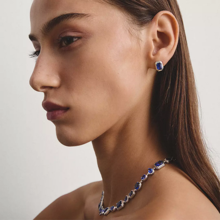A close-up side profile of a woman wearing a matching jewelry set featuring a silver-toned necklace and earring with rectangular blue gemstones.