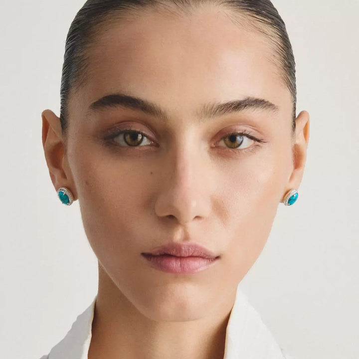 Close-up portrait of a model wearing small round turquoise-blue stud earrings against a light neutral background.
