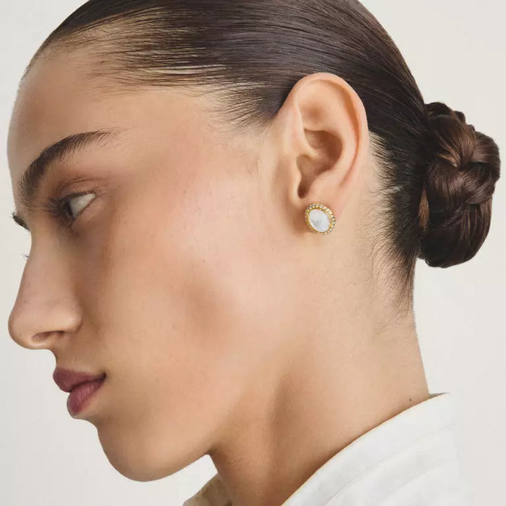 Close-up side profile of a woman wearing a small gold stud earring with a white pearl-like center, against a clean light background.