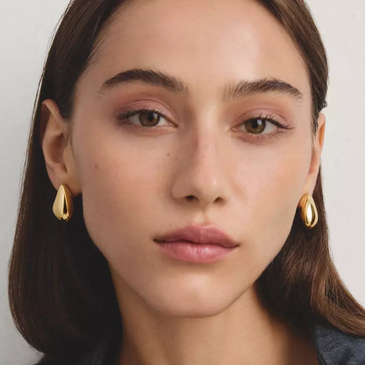 A close-up portrait of a woman wearing small gold teardrop-shaped earrings against a light neutral background.