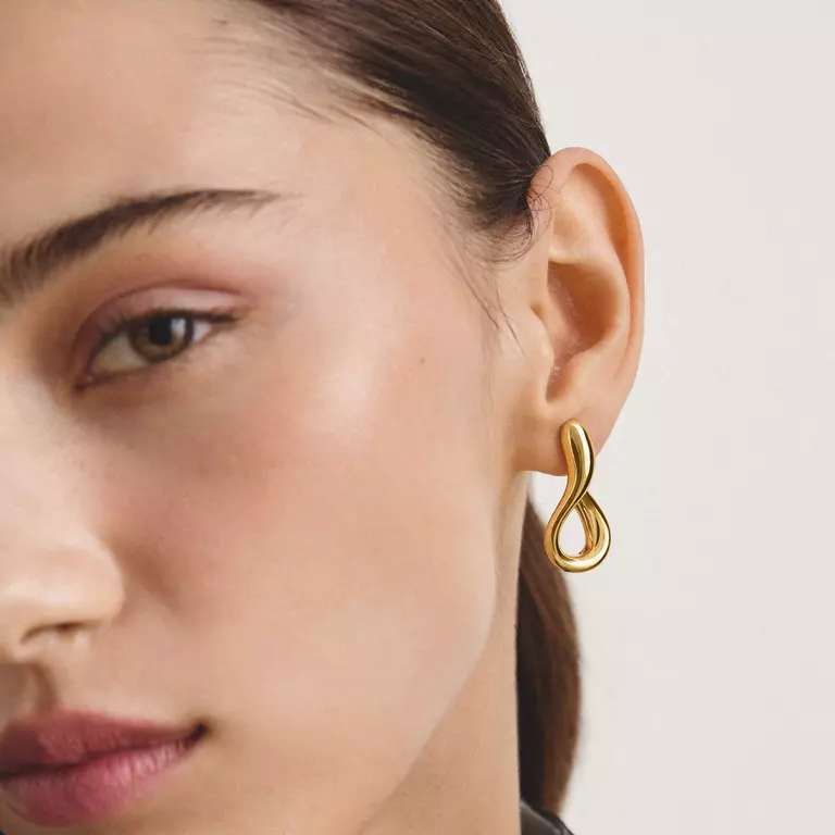 A close-up of a model's ear wearing a polished gold infinity-shaped hoop earring against a clean, light off-white background.