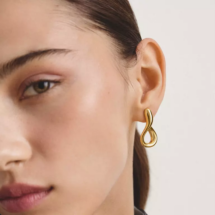 A close-up of a model's ear wearing a polished gold infinity-shaped hoop earring against a clean, light off-white background.