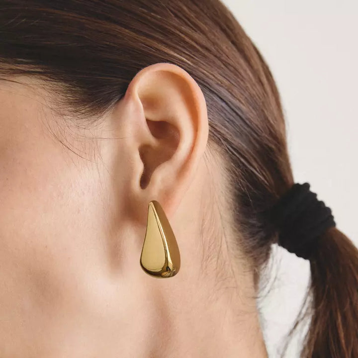 A close-up of a model's ear wearing a polished gold teardrop-shaped stud earring, with her brown hair tied back in a black scrunchie against a light off-white background.
