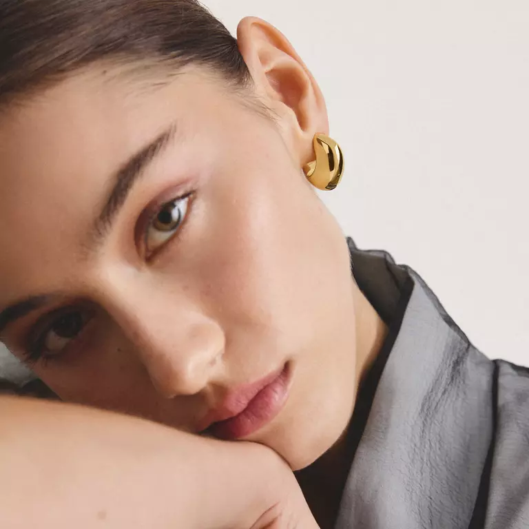 A close-up of a model wearing a gold crescent-shaped hoop earring, resting her head on her hand against a light off-white background.