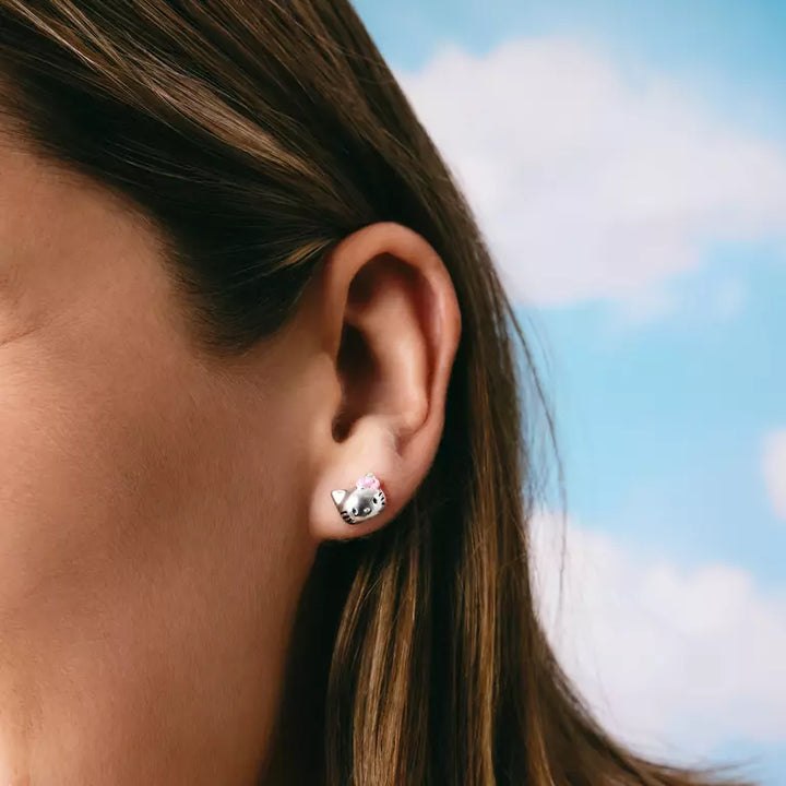 A close-up of a person's ear wearing a small silver stud earring shaped like a cat face with pink inner ears, set against a blurred blue sky background.
