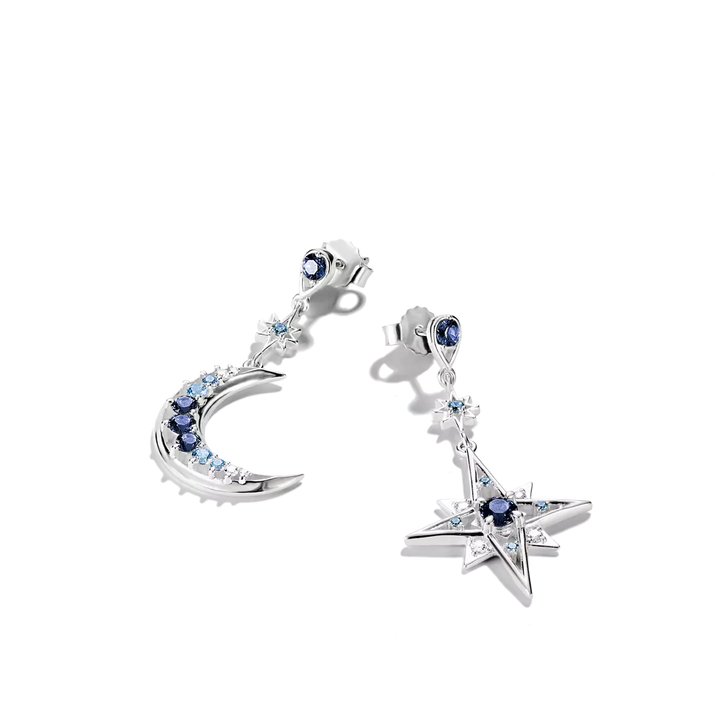 A pair of silver-toned dangle earrings on a plain white background. One earring features a crescent moon shape with blue and clear stones, and the other features a five-pointed star with a blue center stone and clear stones.
