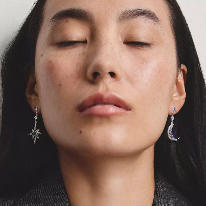 Close-up of a woman wearing delicate silver dangle earrings, one shaped like a star and the other like a crescent moon, both adorned with small blue gemstones.