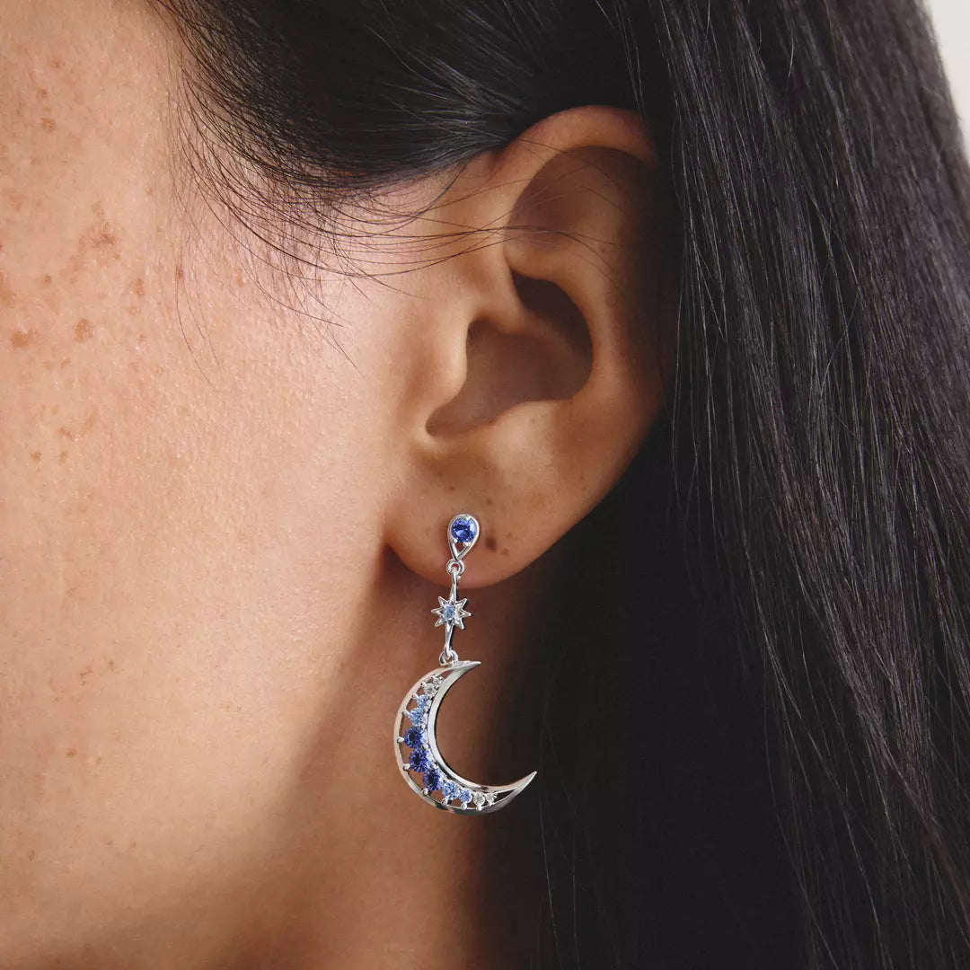A close-up of a person's ear wearing a dangling celestial earring. The earring features a silver crescent moon and star design set with deep blue and clear crystals, shown against the model's skin and dark hair.