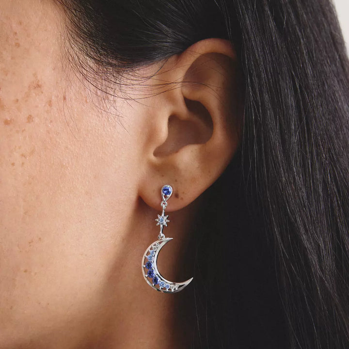 A close-up of a person's ear wearing a dangling celestial earring. The earring features a silver crescent moon and star design set with deep blue and clear crystals, shown against the model's skin and dark hair.