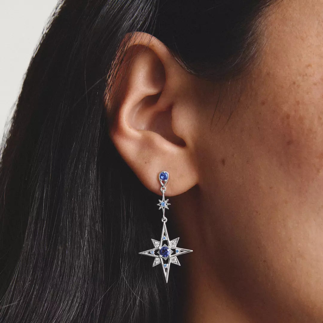Close-up of a woman wearing delicate silver dangle earrings, one shaped like a star and the other like a crescent moon, both adorned with small blue gemstones.