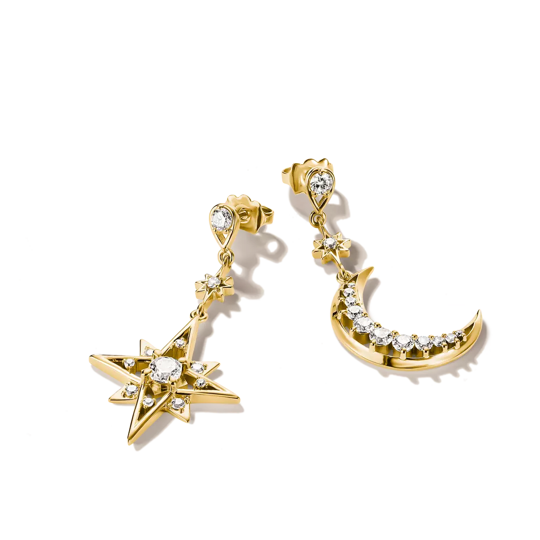 A pair of gold dangle earrings on a clean white background. One earring features a starburst motif with clear gemstones, and the other features a crescent moon with a row of clear gemstones along its curve.