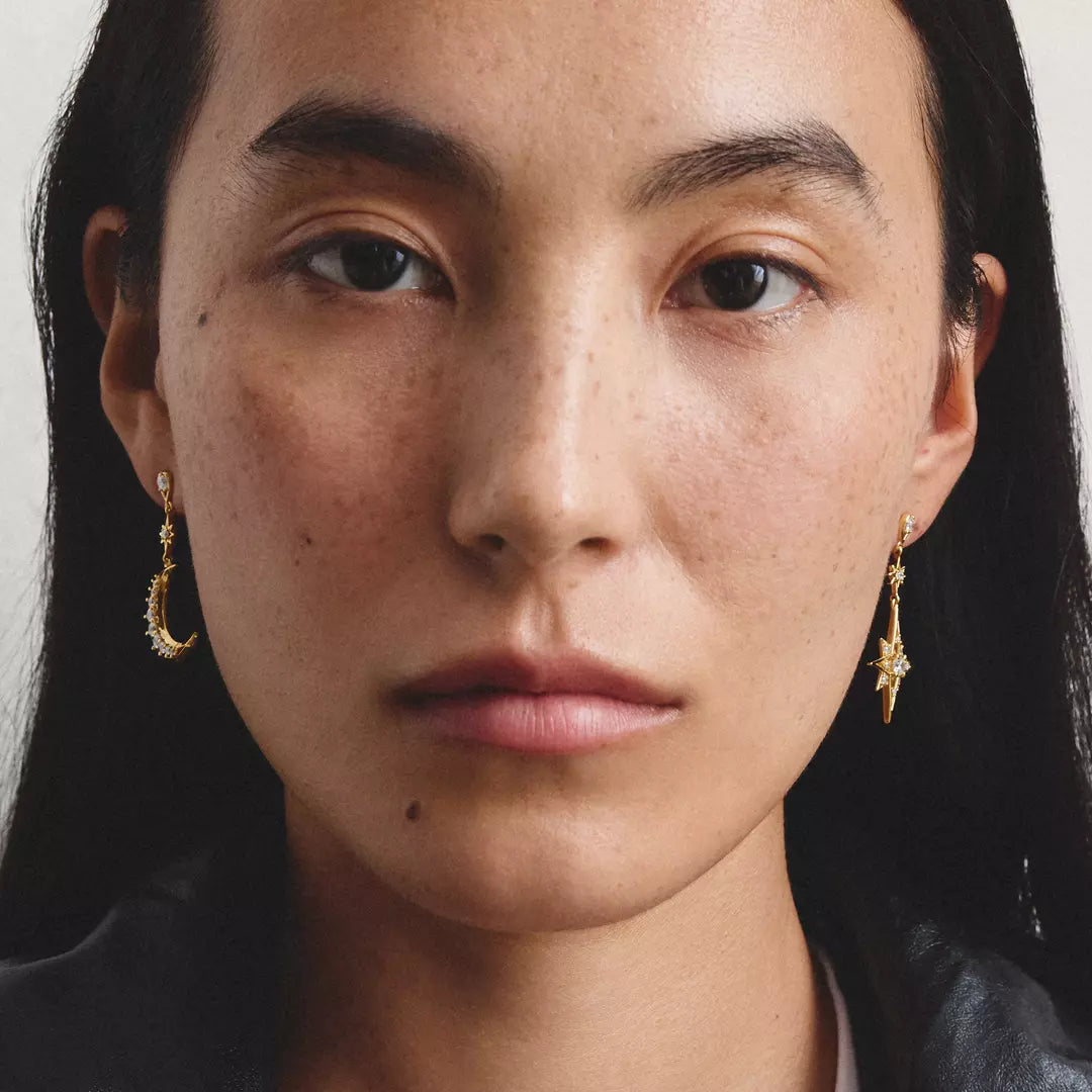 Close-up portrait of a woman wearing small gold crescent moon and star dangle earrings against a plain light background.