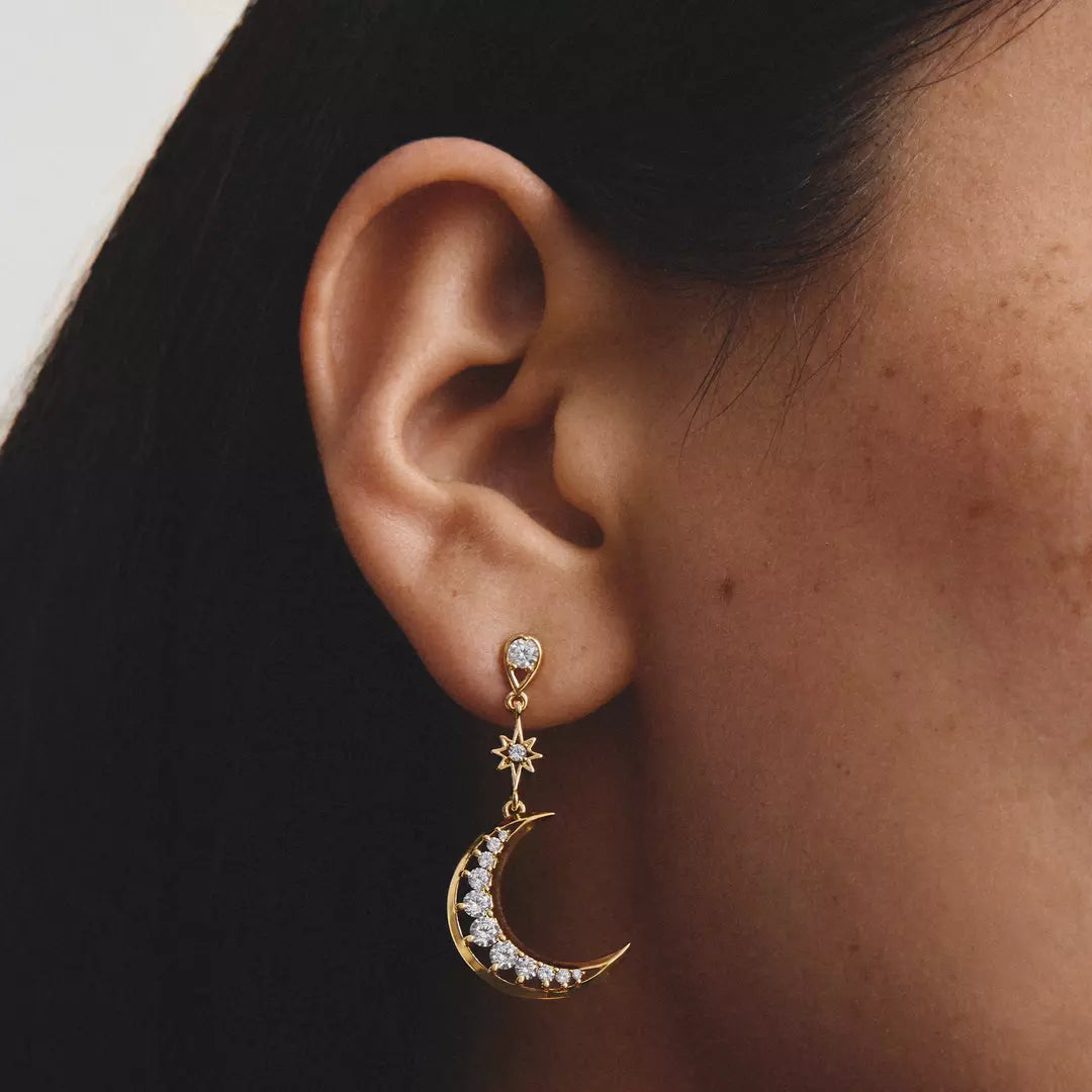 A close-up of a person's ear wearing a gold crescent moon dangle earring adorned with small clear stones, set against dark hair and a light neutral background.