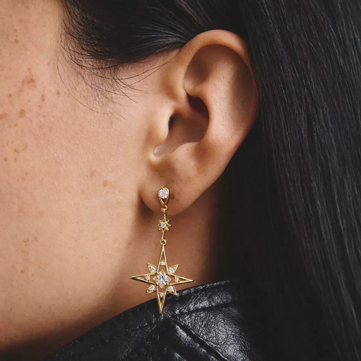 A close-up of a person's ear wearing a gold dangle earring with a starburst design and small clear gemstones.
