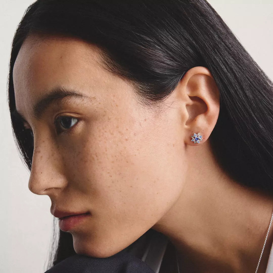 Close-up of a woman wearing a small floral stud earring with blue and white stones on a light neutral background.