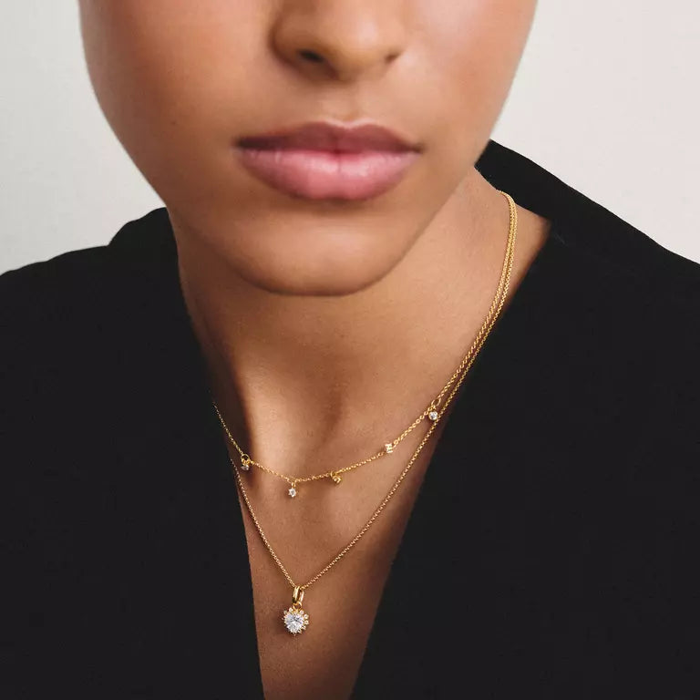 A close-up of a model wearing a delicate gold double-layer necklace. The top layer features tiny gold leaf accents, and the bottom layer has a round clear crystal pendant.