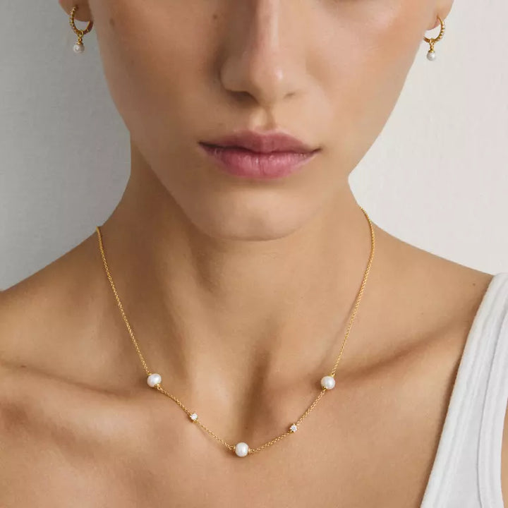 A close-up of a model wearing a delicate gold necklace with five small white pearls spaced along the chain, paired with matching gold hoop earrings.