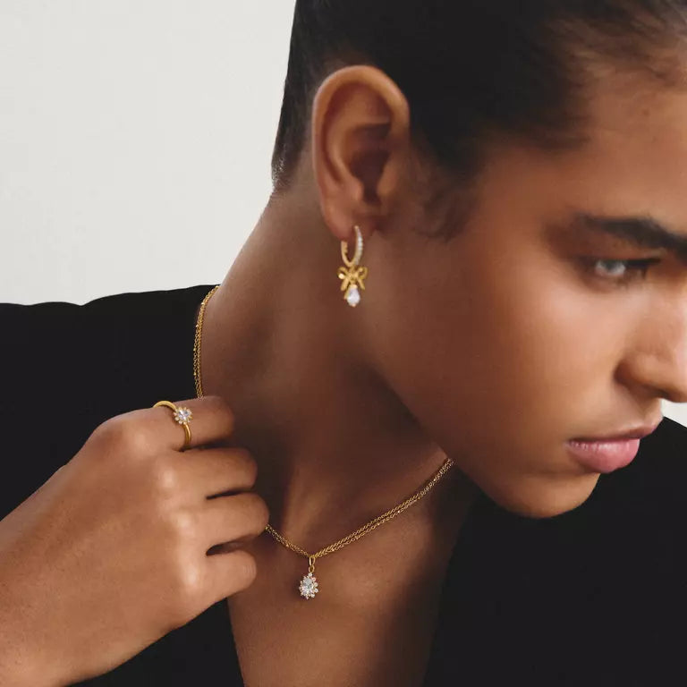 A close-up lifestyle shot of a model wearing a delicate gold necklace with a small clear crystal pendant and a matching gold ring on her finger.