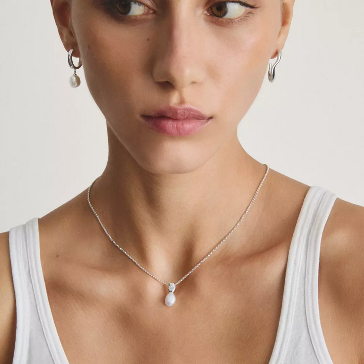 A close-up of a woman wearing a delicate silver chain necklace with a small white teardrop-shaped pearl pendant, paired with matching silver hoop earrings.