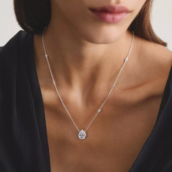 A close-up of a model wearing a delicate silver necklace with a teardrop-shaped clear gemstone pendant, styled with a black top.
