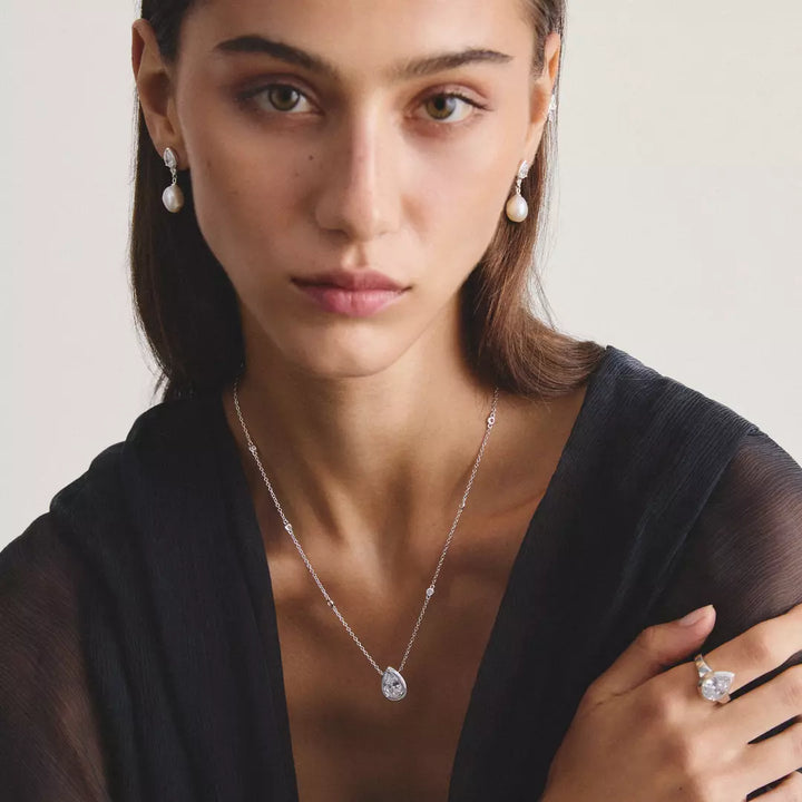 A close-up portrait of a woman wearing a delicate silver necklace with a clear teardrop-shaped pendant, matching pearl drop earrings, and a silver ring with a clear stone, set against a soft light gray background.