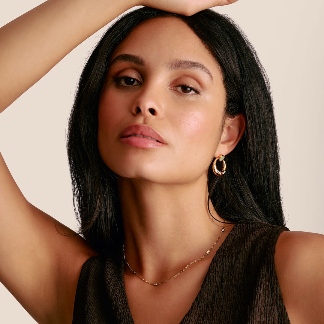 Woman with long dark hair wearing a gold earring and necklace against a beige background