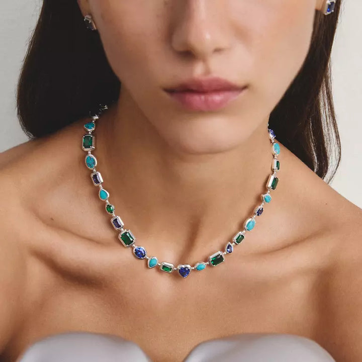 A close-up lifestyle shot of a model wearing a silver-toned necklace adorned with colorful, irregular-cut gemstones in green, turquoise blue, and dark blue, paired with matching stud earrings.