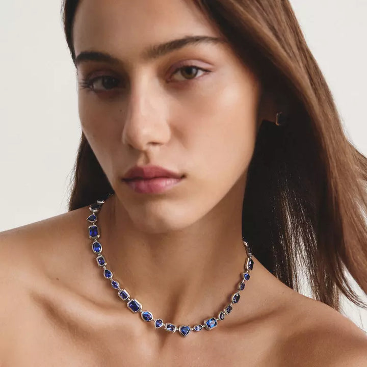 A close-up of a woman wearing a delicate silver-toned necklace set with small oval-cut deep blue gemstones, set against a clean, light off-white background.