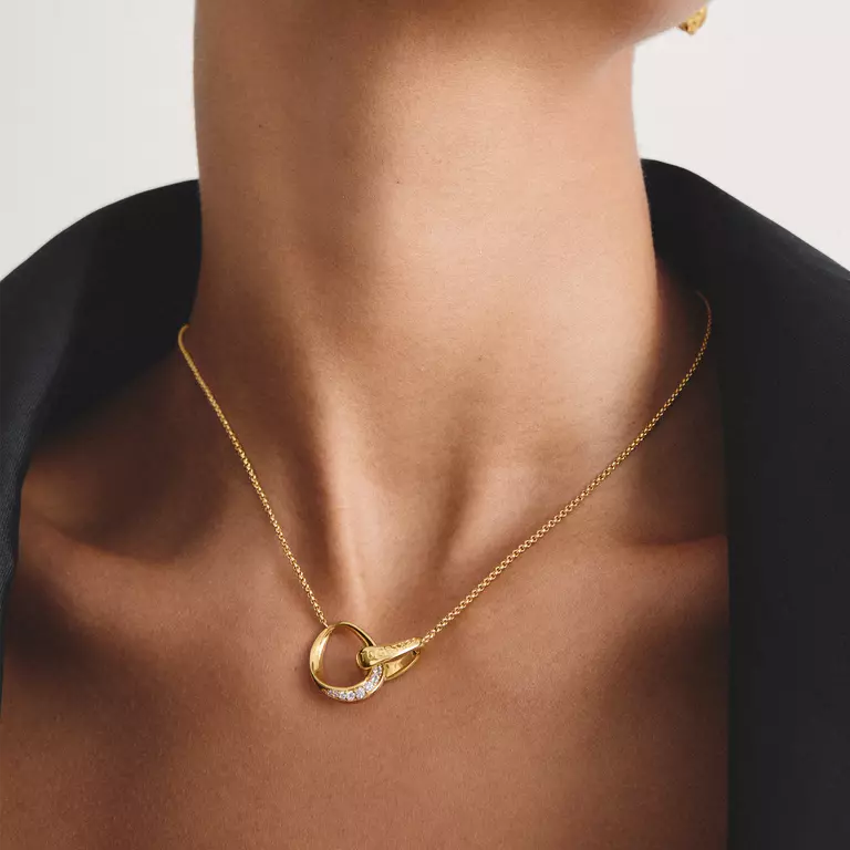 A gold necklace featuring an open interlocking ring pendant with a small row of clear stones on one side and a teardrop-shaped gold element on the other, shown on a white background.