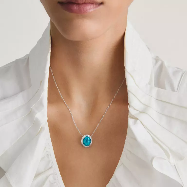 A close-up of a model wearing a delicate silver chain necklace with an oval turquoise pendant, styled with a white ruffled blouse.