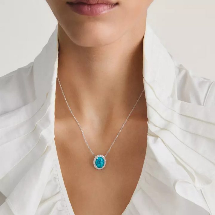 A close-up of a model wearing a delicate silver chain necklace with an oval turquoise pendant, styled with a white ruffled blouse.