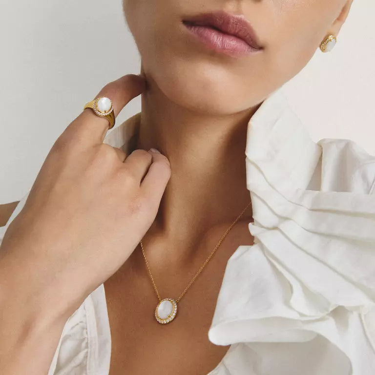 Close-up of a model wearing a delicate gold necklace with a white oval pendant, a matching gold ring, and a gold stud earring, paired with a white ruffled blouse.