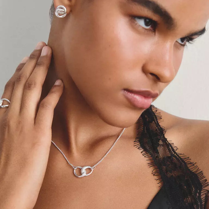 A close-up lifestyle shot of a model wearing a silver double interlocking ring necklace and matching silver stud earrings, styled with a black lace top against a neutral light gray background.