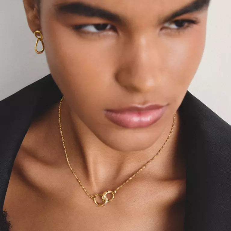 A close-up of a model wearing a delicate gold chain necklace with a double interlocking ring pendant, along with a matching gold teardrop hoop earring, styled with a black blazer against a light gray background.
