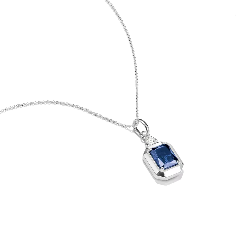 A silver pendant necklace featuring a rectangular emerald-cut blue gemstone, accented by a small round clear stone on the upper left, shown on a clean white background.