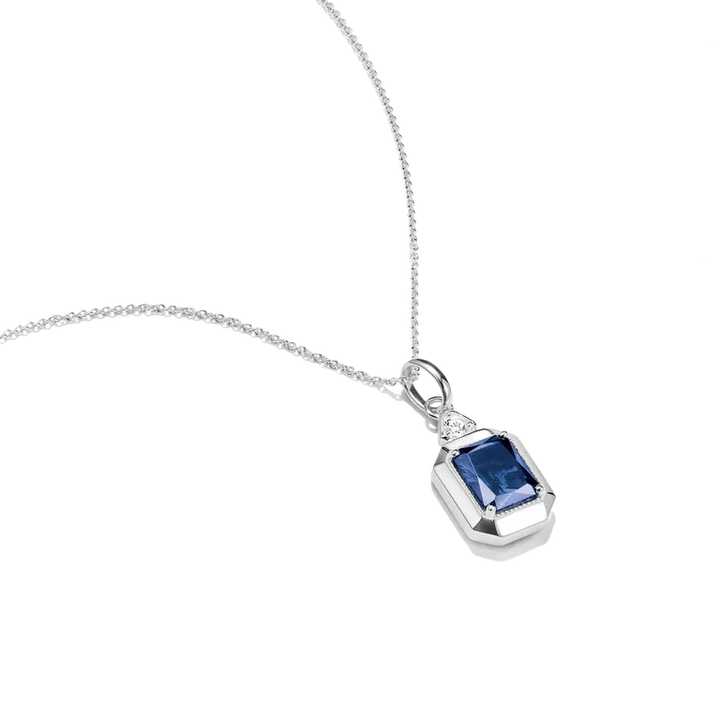A silver pendant necklace featuring a rectangular emerald-cut blue gemstone, accented by a small round clear stone on the upper left, shown on a clean white background.